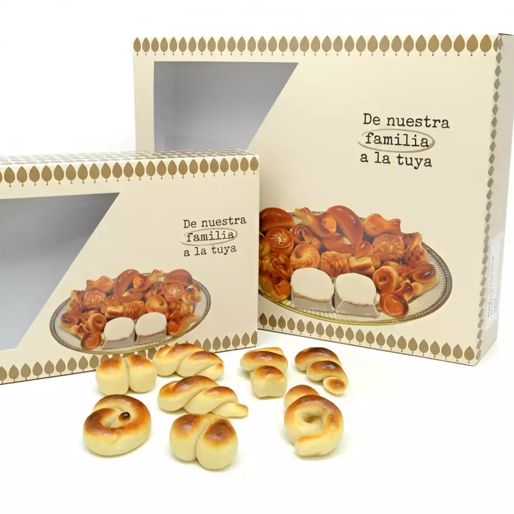 Dos cajas de cartón beige con un patrón de rombos marrones y la frase "De nuestra familia a la tuya". Ambas cajas tienen una ventana transparente que muestra un surtido de dulces de mazapán y almendra. En primer plano, varios dulces de mazapán en espiral y con forma de lazo están esparcidos.